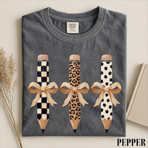 A gray t-shirt with a graphic design of three pencils with bows and leopard print patterns.