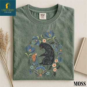 a green t-shirt with a colorful floral and animal design, including a black bear, flowers, and other elements.