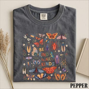 A gray t-shirt with a colorful graphic design featuring various insects and flowers, along with the text "Be kind to all kinds".
