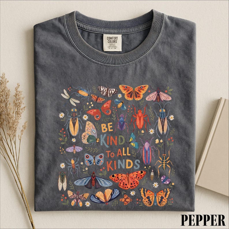 A gray t-shirt with a colorful graphic design featuring various insects and flowers, along with the text "Be kind to all kinds".