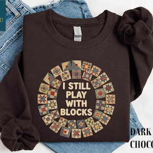 May include: Dark chocolate-colored sweatshirt with the text "I STILL PLAY WITH BLOCKS" in a circular design. The design features a quilt block pattern in various colors. The words "DARK CHOCOLATE" are printed below the sweatshirt.