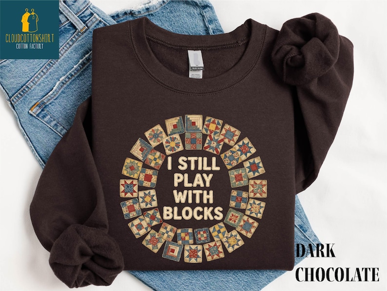 May include: Dark chocolate-colored sweatshirt with the text "I STILL PLAY WITH BLOCKS" in a circular design. The design features a quilt block pattern in various colors. The words "DARK CHOCOLATE" are printed below the sweatshirt.