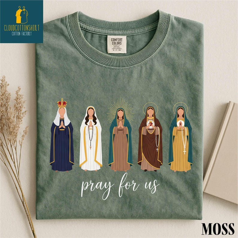May include: A moss green t-shirt featuring a graphic of five religious figures with halos and crowns. The words "pray for us" are printed in white script below the figures. The shirt has a Comfort Colors tag.