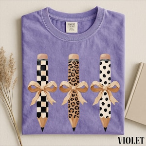 a purple t-shirt with three pencils with different designs and bows on them, placed on a white surface.