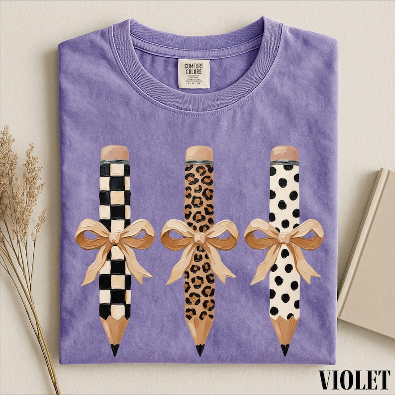 a purple t-shirt with three pencils with different designs and bows on them, placed on a white surface.