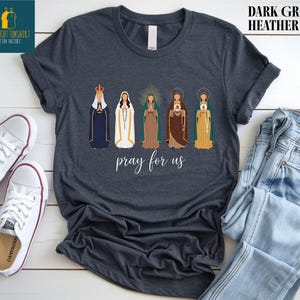 May include: Dark gray heather t-shirt featuring an illustration of five religious figures in various robes and crowns. The phrase "pray for us" is written in cursive below the figures. The shirt is displayed with white sneakers and blue jeans.