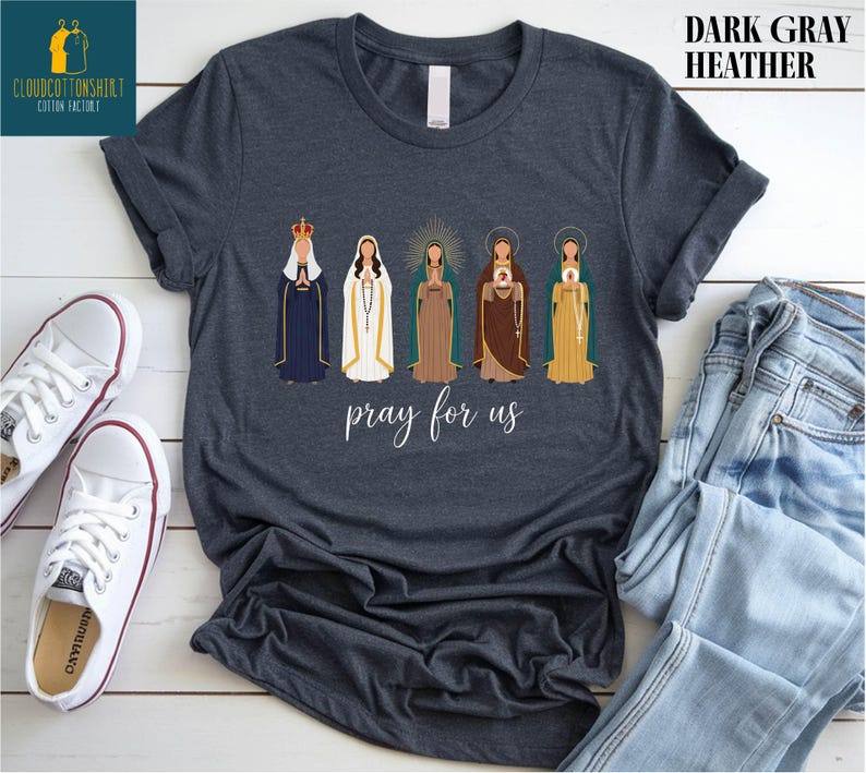 May include: Dark gray heather t-shirt featuring an illustration of five religious figures in various robes and crowns. The phrase "pray for us" is written in cursive below the figures. The shirt is displayed with white sneakers and blue jeans.