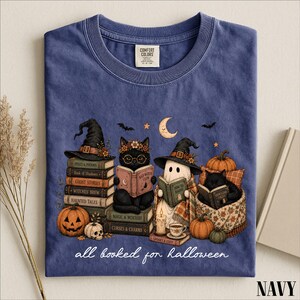 A navy blue t-shirt with a Halloween-themed graphic design depicting a witch, cat, ghost, pumpkin, and books, along with the text "all baked for Halloween".