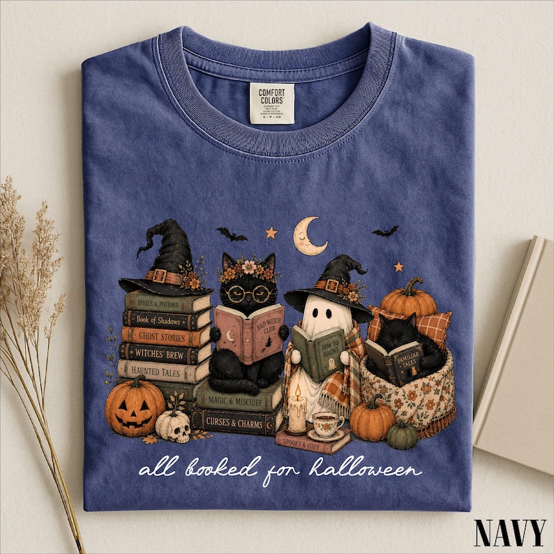 A navy blue t-shirt with a Halloween-themed graphic design depicting a witch, cat, ghost, pumpkin, and books, along with the text "all baked for Halloween".