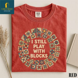 May include: Red t-shirt with a round graphic of quilt blocks and the text "I STILL PLAY WITH BLOCKS". The shirt has a crew neck and short sleeves. The shirt is folded and has a tag that says "Comfort Colors".