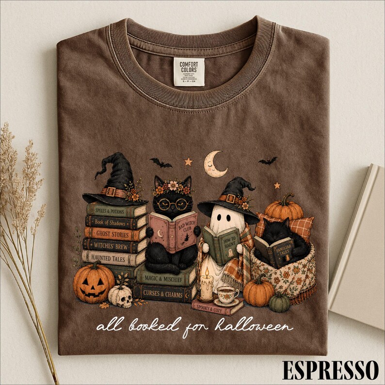 A t-shirt with a Halloween-themed design featuring cats dressed as witches and a ghost reading books, surrounded by pumpkins, gourds, and other Halloween-related items.