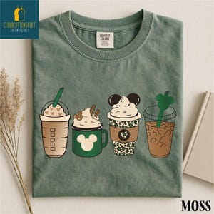 May include: Moss green t-shirt featuring four illustrated coffee drinks. The drinks include a Starbucks-style cup with hearts, a green mug with a Mickey Mouse design, a cup with a leopard print, and a clear cup with a green straw.