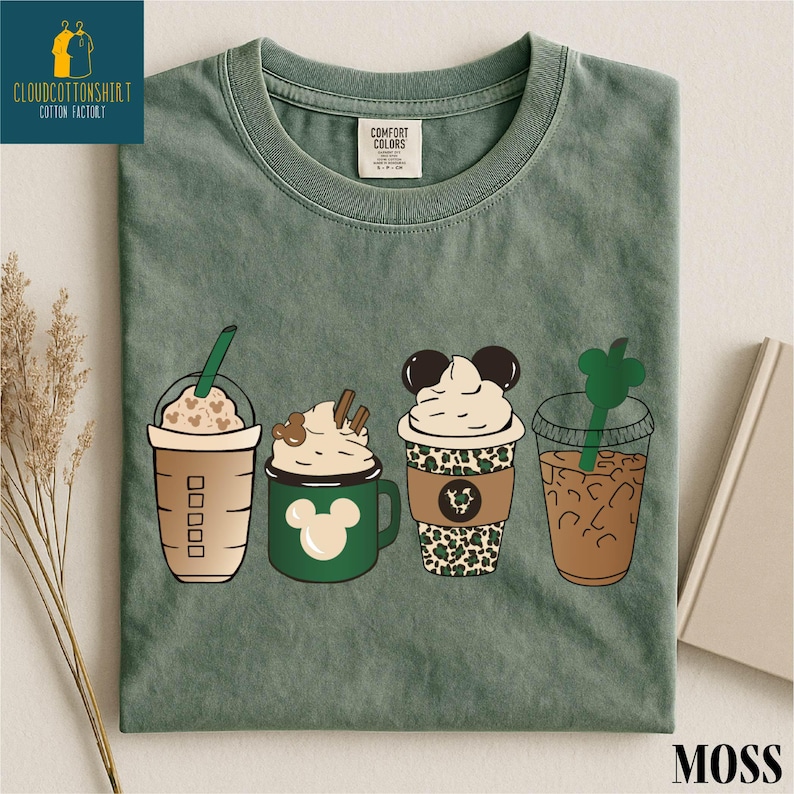 May include: Moss green t-shirt featuring four illustrated coffee drinks. The drinks include a Starbucks-style cup with hearts, a green mug with a Mickey Mouse design, a cup with a leopard print, and a clear cup with a green straw.