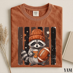 May include: A rust-colored t-shirt featuring a graphic of a raccoon wearing an orange knit beanie, holding a coffee cup with a pumpkin icon and a brown football. The design is set against a background of vertical dark stripes and small stars.