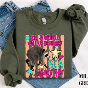 A military green sweatshirt with a graphic design featuring a raccoon and the text "In a world full of ordinary, be a jimothy"