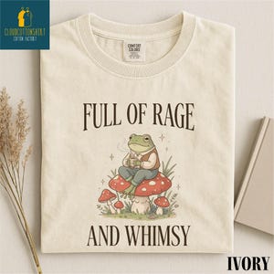 a white t-shirt with a printed design depicting a frog sitting on a mushroom, surrounded by mushrooms and plants. The text on the t-shirt reads "FULL OF RAGE AND WHIMSY".