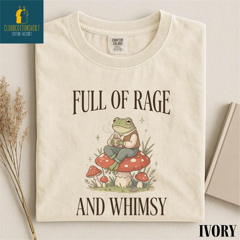 a white t-shirt with a printed design depicting a frog sitting on a mushroom, surrounded by mushrooms and plants. The text on the t-shirt reads "FULL OF RAGE AND WHIMSY".