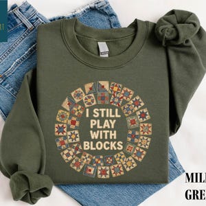 May include: Military green sweatshirt with the text "I STILL PLAY WITH BLOCKS" in white. The text is surrounded by a circular design of colorful quilt blocks. The sweatshirt is folded on top of blue jeans.