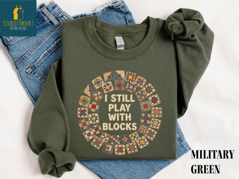 May include: Military green sweatshirt with the text "I STILL PLAY WITH BLOCKS" in white. The text is surrounded by a circular design of colorful quilt blocks. The sweatshirt is folded on top of blue jeans.