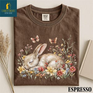 May include: Espresso-colored t-shirt featuring a sleeping bunny surrounded by colorful flowers and butterflies. The shirt has a Comfort Colors tag and the text "ESPRESSO" at the bottom. The Cloud Cotton Shirt logo is in the upper left corner.
