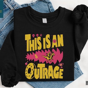 A black t-shirt with a graphic design featuring the text "THIS IS AN OUTRAGE" and a dinosaur-like creature.