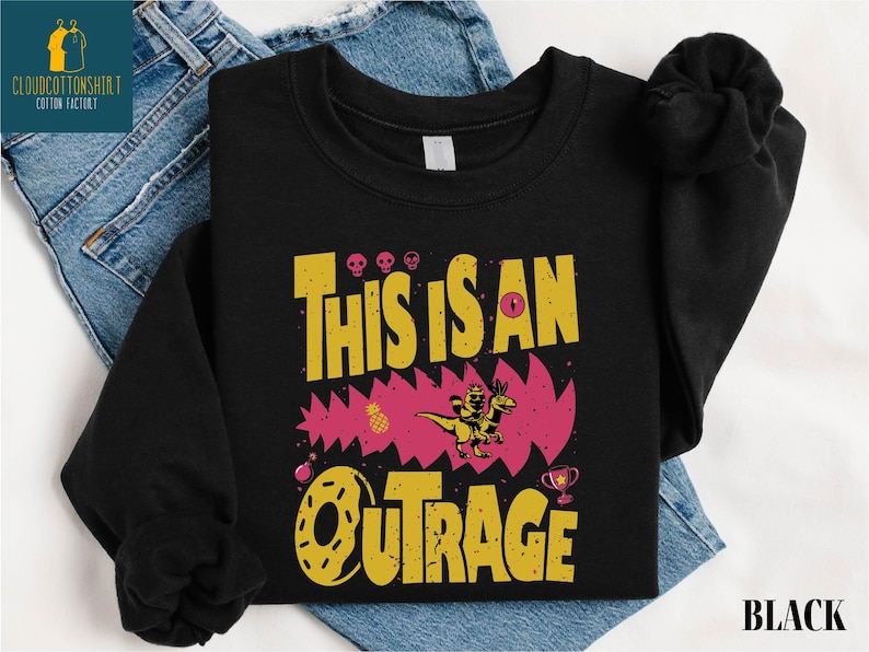 A black t-shirt with a graphic design featuring the text "THIS IS AN OUTRAGE" and a dinosaur-like creature.