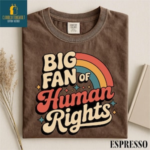 May include: Espresso-colored t-shirt with the text "BIG FAN of Human Rights" in a retro font, featuring a rainbow design. The shirt has a crew neck and a Comfort Colors tag.