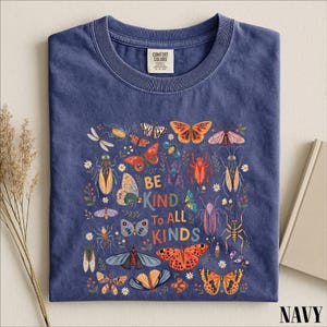 A navy blue t-shirt with a colorful graphic design featuring various insects and butterflies, along with the text "Be kind to all kinds".