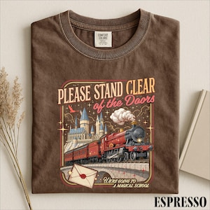 May include: A brown short-sleeve t-shirt featuring a graphic of a red steam train crossing a stone bridge toward a castle. The text reads: PLEASE STAND CLEAR of the Doors and WE'RE GOING TO A MAGICAL SCHOOL, with an envelope illustration.