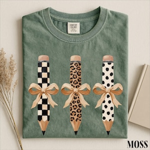 A green t-shirt with three pencils with different designs and bows on them, along with some dried flowers and a book.
