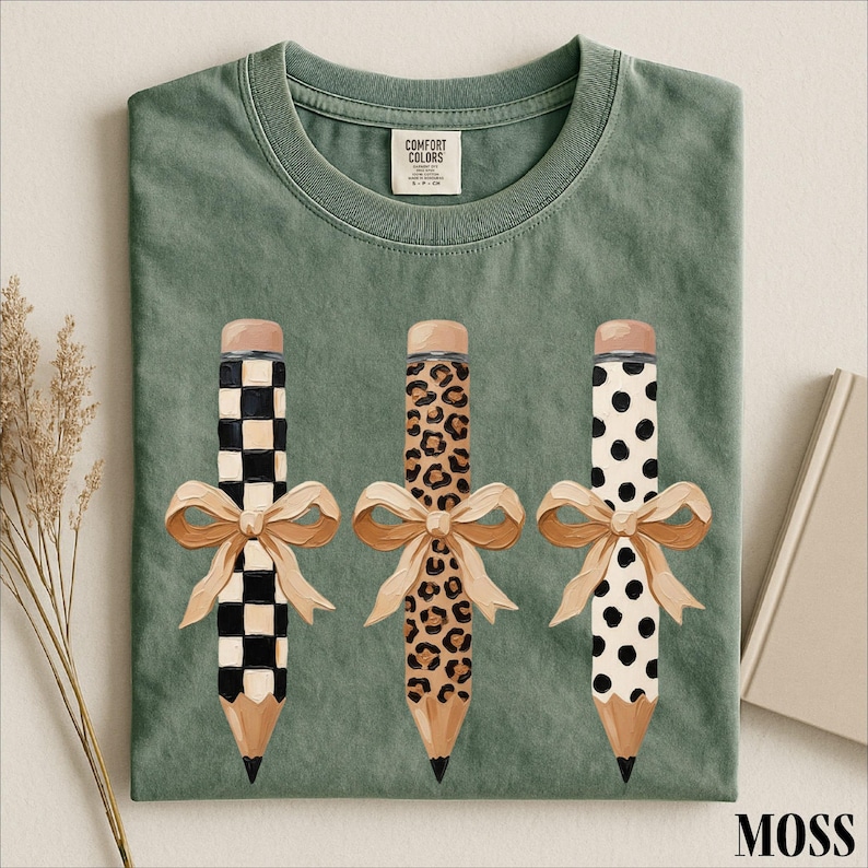 A green t-shirt with three pencils with different designs and bows on them, along with some dried flowers and a book.