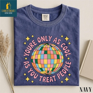 May include: Navy blue t-shirt with a colorful disco ball graphic and the text "YOU'RE ONLY AS COOL AS YOU TREAT PEOPLE." The shirt also has yellow star accents. The shirt is folded on a neutral surface.