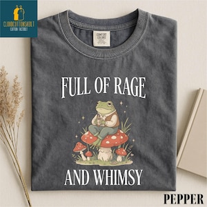 a gray t-shirt with a whimsical design of a frog sitting on a mushroom, surrounded by mushrooms and other natural elements. The text on the t-shirt reads "FULL OF RAGE AND WHIMSY".