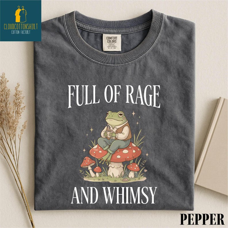 a gray t-shirt with a whimsical design of a frog sitting on a mushroom, surrounded by mushrooms and other natural elements. The text on the t-shirt reads "FULL OF RAGE AND WHIMSY".