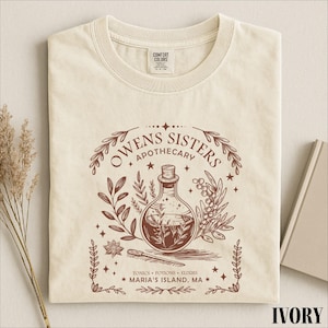 May include: An ivory t-shirt featuring a brown graphic of a potion bottle, botanical sprigs, and the text "Owens Sisters Apothecary" along with "Tonics, Potions, Elixirs" and "Maria's Island, MA". The shirt has a crew neckline and a Comfort Colors label.