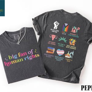 May include: Two dark gray t-shirts. One shirt has the text "big fan of human rights" in colorful letters. The other shirt has various graphics and text including "Black Lives Matter" and "Feminism is for everyone".