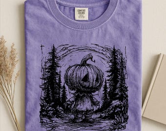 May include: A violet short-sleeve t-shirt featuring a black ink-style graphic of a pumpkin-headed figure standing in a forest of tall pine trees. The shirt has a crew neckline and a Comfort Colors brand label printed on the inside of the collar.