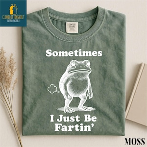 May include: A moss green t-shirt with the text "Sometimes I Just Be Fartin'" and a cartoon frog illustration. The shirt has a crew neck and a Comfort Colors tag.