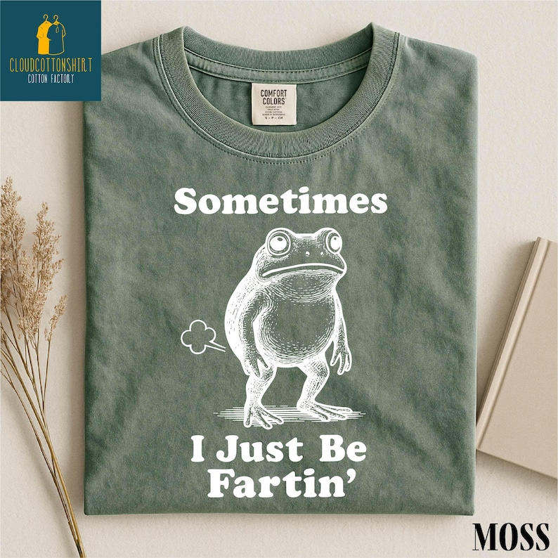 May include: A moss green t-shirt with the text "Sometimes I Just Be Fartin'" and a cartoon frog illustration. The shirt has a crew neck and a Comfort Colors tag.