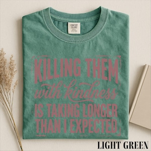A green t-shirt with a printed message that reads "Killing them with kindness is taking longer than I expected" in pink text.