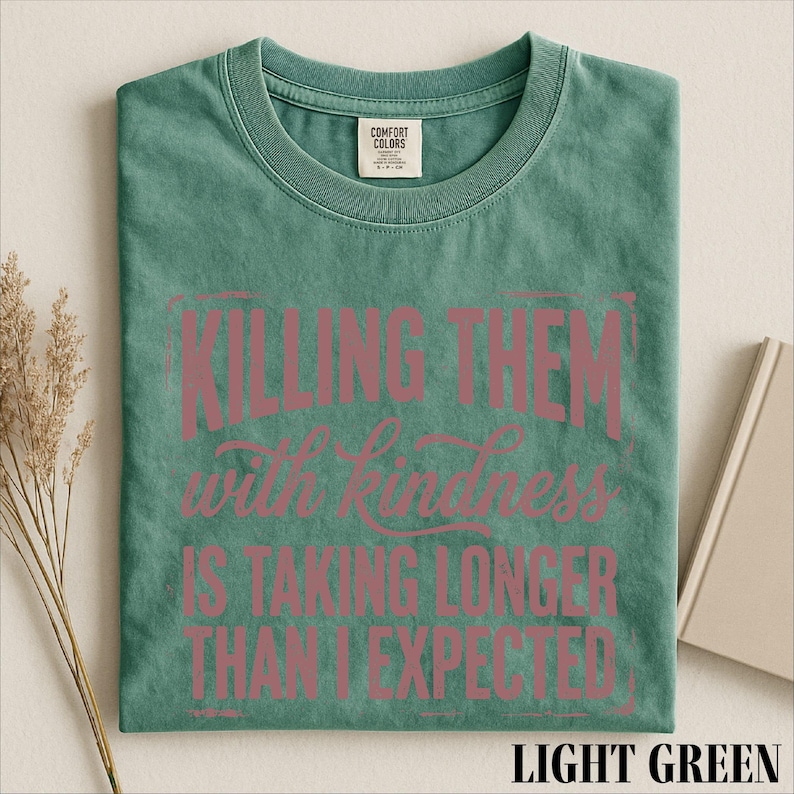 A green t-shirt with a printed message that reads "Killing them with kindness is taking longer than I expected" in pink text.