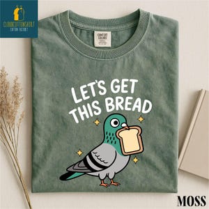 May include: Moss green t-shirt with the text "LET'S GET THIS BREAD" and a cartoon pigeon holding a slice of bread. The shirt has a crew neck and short sleeves. The shirt is made of a soft, comfortable material.