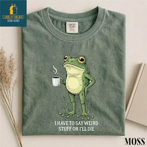 A green t-shirt with a cartoon frog holding a cup of coffee and the text "I HAVE TO SAY WEIRD STUFF OR I'll die" printed on it.