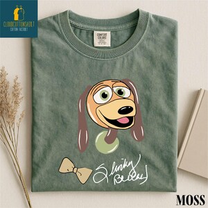 A green t-shirt with a cartoon dog design and a bow tie, along with a label that says "Comfort Color" and "Moss".