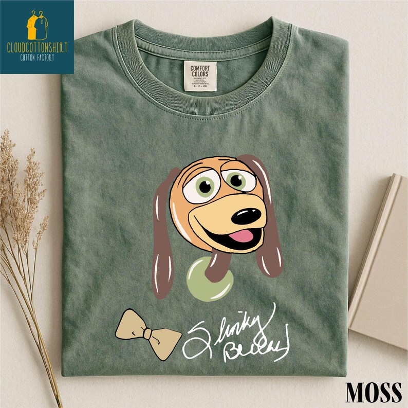 A green t-shirt with a cartoon dog design and a bow tie, along with a label that says "Comfort Color" and "Moss".