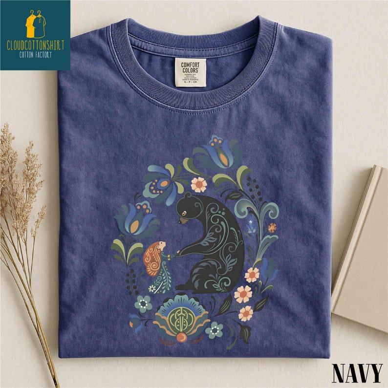 A navy blue t-shirt with a colorful floral and animal design, including a bear, flowers, and other elements, is displayed on a white surface.