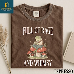 a brown t-shirt with a whimsical design of a frog sitting on a mushroom, surrounded by mushrooms and other natural elements. The text on the t-shirt reads "FULL OF RAGE AND WHIMSY" and "ESPRESSO".