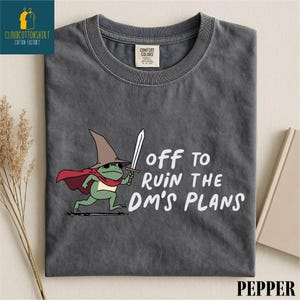 May include: A dark gray t-shirt featuring a cartoon frog wearing a wizard hat and cape, holding a sword. The text "off to ruin the DM's plans" is printed in white. The shirt has a Comfort Colors tag.