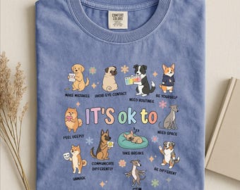 May include: A blue jean colored t-shirt featuring a graphic of ten dogs with the text "IT'S OK to" and various phrases including "MAKE MISTAKES," "AVOID EYE CONTACT," "NEED ROUTINES," "BE YOURSELF," "FEEL DEEPLY," "NEED SPACE," "UNMASK," "COMMUNICATE DIFFERENTLY," "TAKE BREAKS," "BE DIFFERENT," and "STIM."