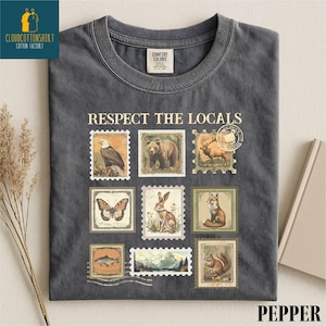 May include: Dark gray t-shirt with the text "RESPECT THE LOCALS" above a collage of vintage-style postage stamps featuring wildlife. Stamps include an eagle, bear, deer, butterfly, rabbit, fox, fish, mountains, and a squirrel.